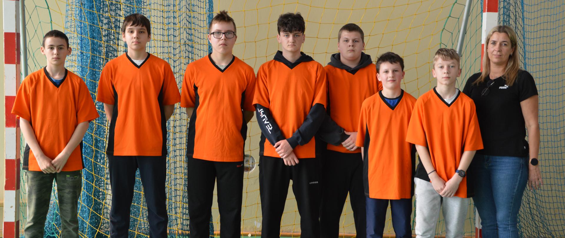 Siedmiu chłopców stojących w rzędzie na tle bramki do piłki nożnej i ubranych w pomarańczowe koszulki. Obok nich z prawej strony stoi opiekunka drużyny.