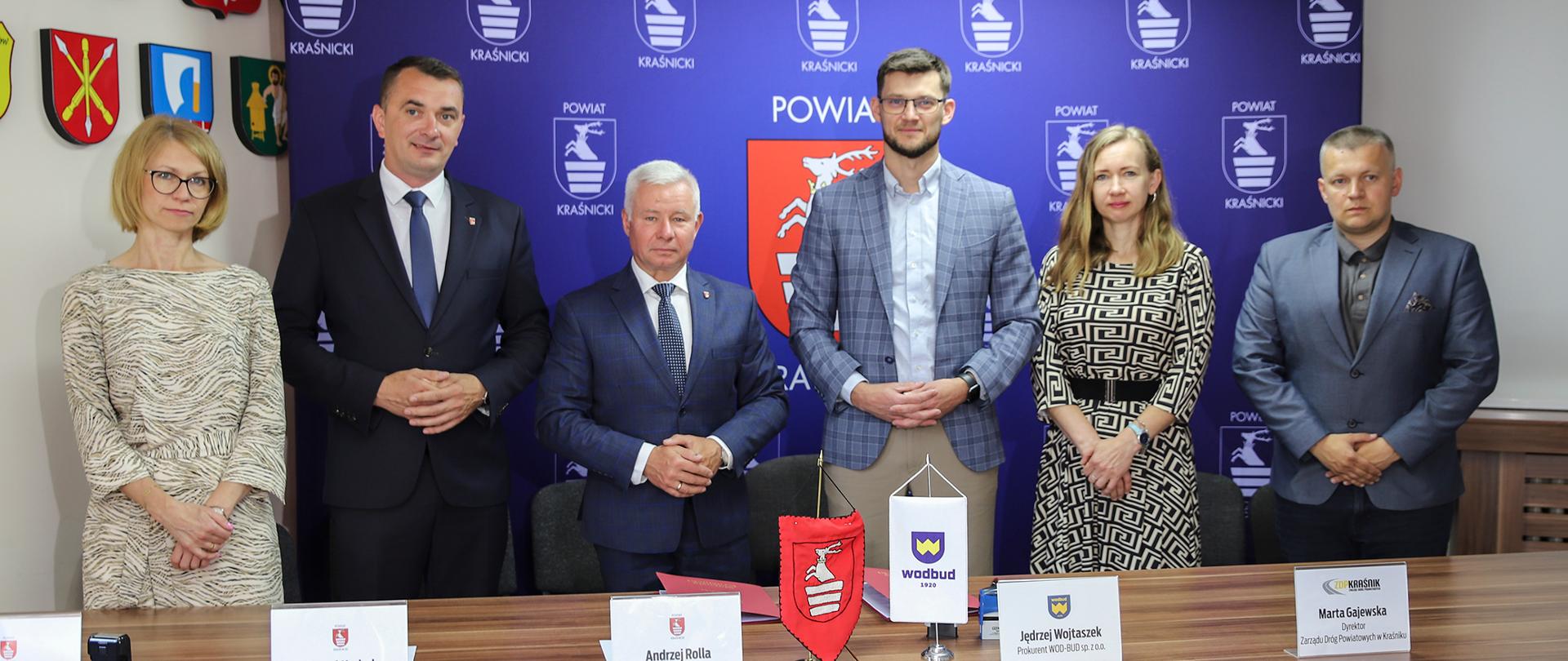 Pamiątkowe zdjęcie z podpisania umowy na budowę drogi. Zebrani stoją w rzędzie na tle niebieskiej ścianki z wizerunkiem herbu powiatu.