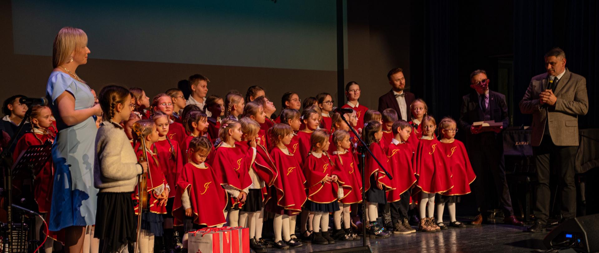 Na scenie Sali Widowiskowej stoi liczna grupa dzieci w czerwonych pelerynkach ze złotymi lamówkami, tworząca chór Talentowni Jana Pawła II. Obok nich stoją dorośli prowadzący wydarzenie. Po prawej stronie znajduje się mężczyzna w szarej marynarce przemawiający do mikrofonu, a obok niego dwóch prowadzących w garniturach. Po lewej stronie stoi kobieta w jasnoniebieskiej sukience. W tle widoczne jest duże logo Centrum Kultury, Sportu i Turystyki w Kalwarii Zebrzydowskiej. Cała grupa oświetlona jest scenicznym światłem, a atmosfera zdjęcia oddaje uroczysty i radosny charakter wydarzenia.