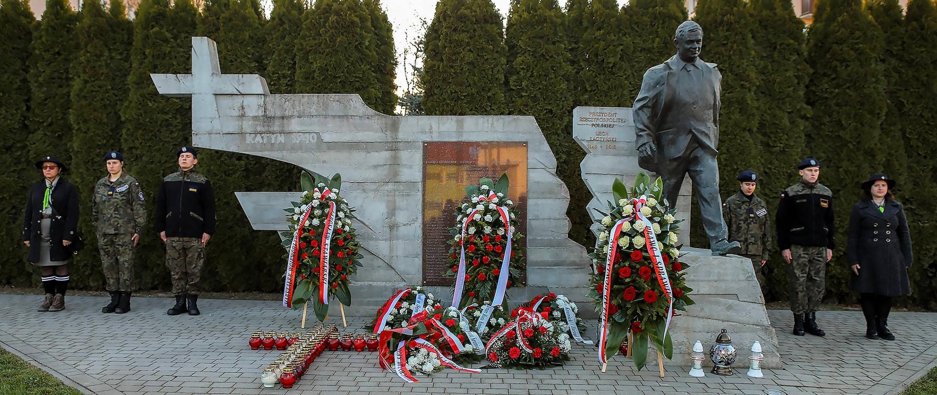 Szerokie ujęcie pomnika upamiętniającego Prezydenta RP Lecha Kaczyńskiego oraz Ofiary katastrofy smoleńskiej, znajdującego się na wybrukowanym placu. W centralnej części widać betonową ścianę o nieregularnych, poszarpanych kształtach, przypominającą statecznik samolotu. Po lewej stronie ściany widnieje napis „KATYŃ 1940” oraz znak krzyża. Po prawej stronie, na podwyższeniu, stoi realistyczny, odlany z brązu posąg Lecha Kaczyńskiego w marszu. Przed pomnikiem złożone są liczne wieńce w barwach narodowych (biało-czerwone kwiaty i szarfy) oraz płonące znicze ułożone w kształt krzyża. Po obu stronach pomnika stoi warta honorowa. Po lewej i po prawej stronie: dwie osoby w mundurach oraz kobieta w stroju harcerskim.