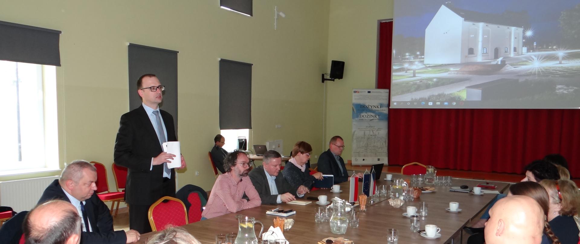 Wystąpienie wójta Grzegorza Sikorskiego. Jedna osoba stoi, reszta siedzi przy dużym stole. W tle na ekranie wyświetlana jest prezentacja, widoczny Dom Przyrodnika w nocnej iluminacji