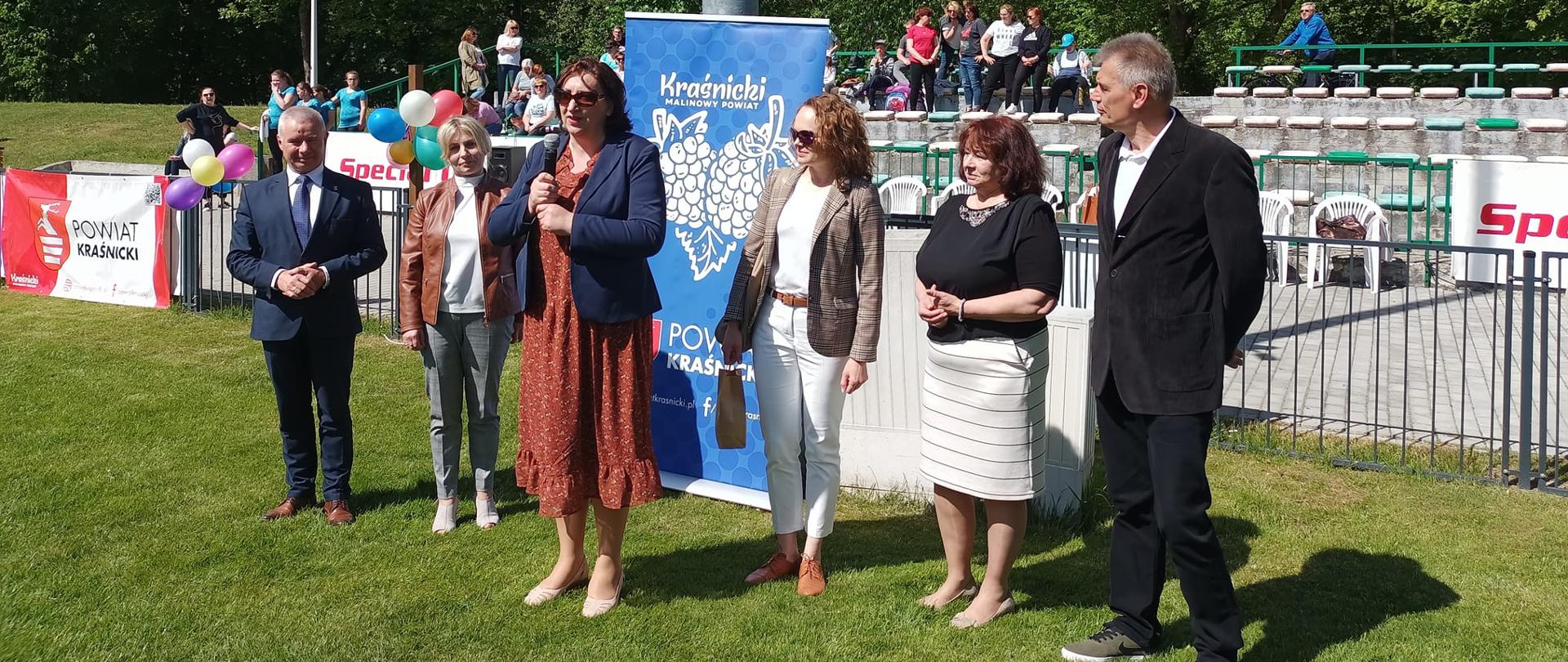 Zdjęcie przedstawia piątkę gości stojących w szeregu na zielonej murawie stadionu podczas uroczystego otwarcia turnieju. Od lewej stoją: Andrzej Rolla - Starosta Kraśnicki, Anna Zdolska - wicedyrektor Specjalnego Ośrodka Szkolno - Wychowawczego w Olbięcinie, Anna Niekra - dyrektor Zespołu Szkół Specjalnych w Kraśniku, Krystyna Krotkiewicz - dyrektor Zespołu Placówek Oświatowych nr 1 w Kraśniku oraz Krystian Wójcik – dyrektor Oddziału Regionalnego Olimpiady Specjalne Polska. Pani Anna Niekra trzyma w rękach mikrofon i przemawia. Z tyłu na trybunach znajdują się ludzie.