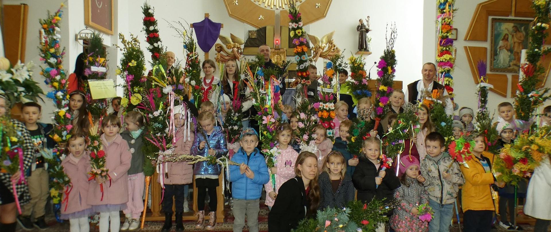 Zdjęcie przedstawia uczestników konkursu Palm Wielkanocnych pozujących do zdjęć na tle ołtarza w kościele parafialnym w Sieniawie.