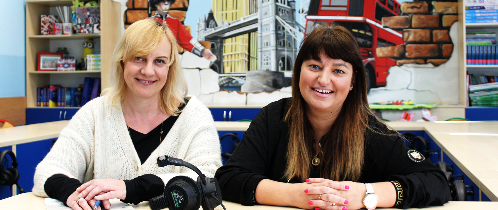 Dwie kobiety siedzą przy stole w klasie języka angielskiego. Przed nimi leżą podręczniki, słuchawki i długopisy. W tle kolorowy mural przedstawiający symbole Londynu – Big Ben, czerwony autobus, strażnika królewskiego i flagę Wielkiej Brytanii