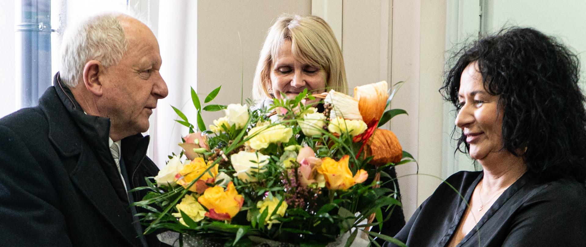 Mężczyzna z lewej strony ubrany w czarny płaszcz przekazuje kobiecie po lewej stronie kosz róż białych, żółtych, herbacianych, w tle stoi trzecia kobieta