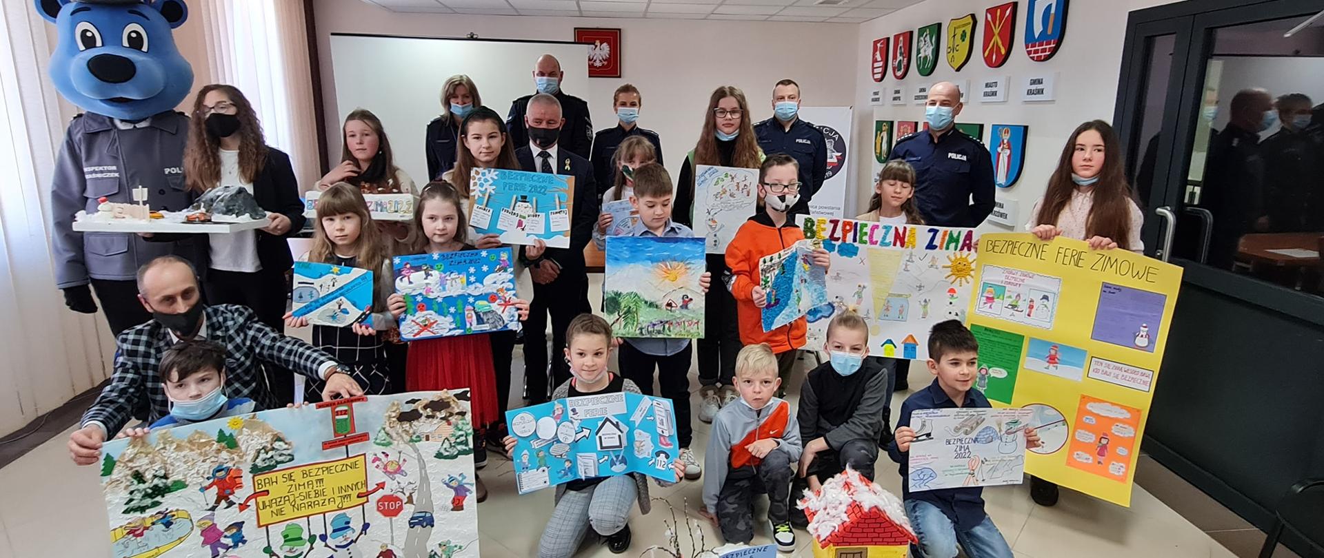 Grupowe zdjęcie laureatów konkursu "Bezpieczna Zima 2022". Dzieci trzymają w ręku swoje konkursowe prace. Po lewej stronie zdjęcia stoi maskotka -miś inspektor Kraśniczek. Za laureatami konkursu stoją: mł.insp. Jacek Figel, Starosta Kraśnicki Andrzej Rolla, podinsp. Dorota Węcławska-Baran, kom. Bartosz Stańsko.