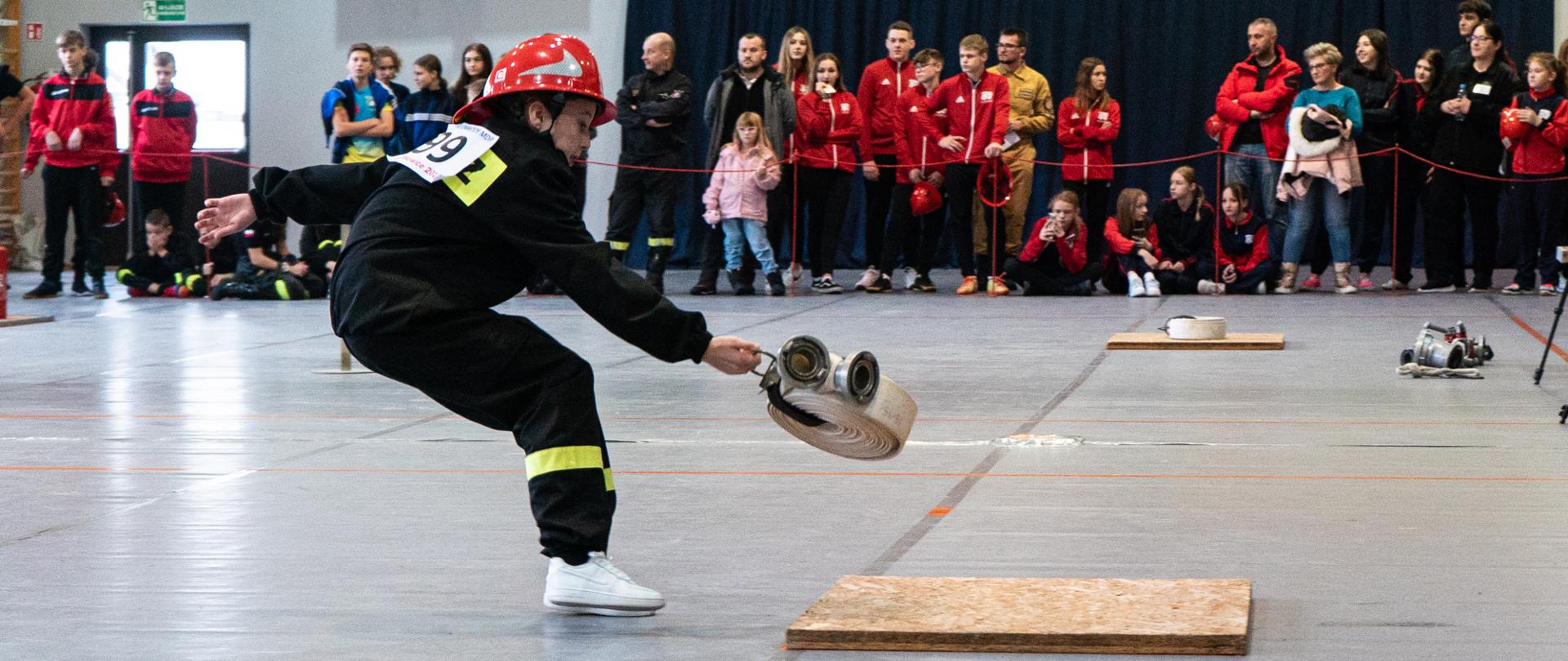 Strażak w czasie zawodów ubrany w rynsztunek strażacki wykonuje rzut zwiniętym wężem strażackim, w tle grupa młodzieży oraz dorosłych kibicujących w zawodach.