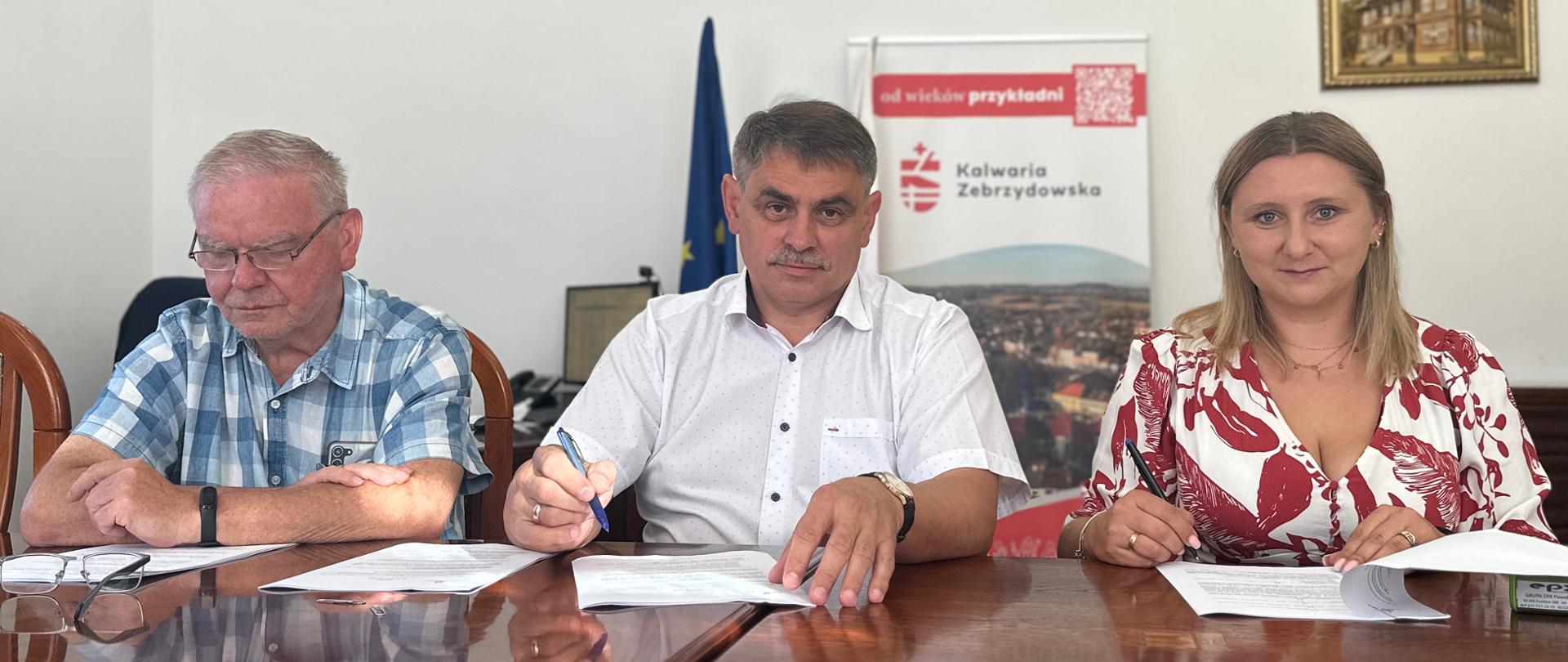 Trzy osoby siedzą przy stole w sali urzędu i podpisują dokumenty. W tle widoczne są godło Polski, flaga Unii Europejskiej oraz baner promocyjny Kalwarii Zebrzydowskiej.