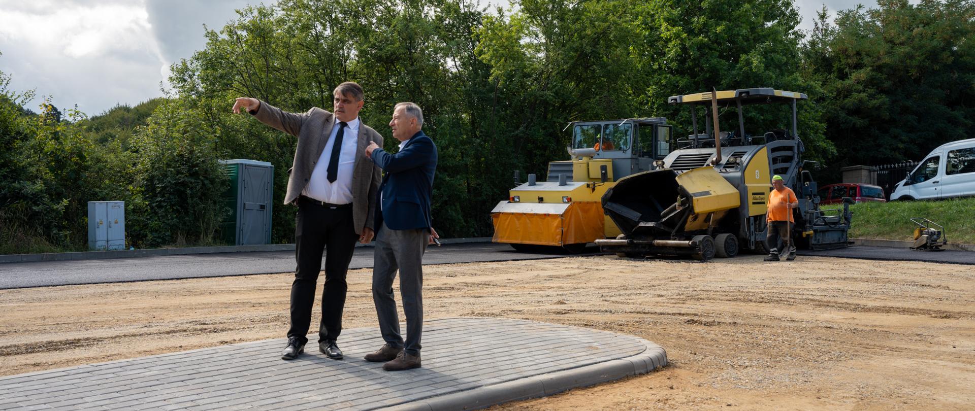 Dwóch mężczyzn w garniturach stoi na nowo ułożonym chodniku i wskazuje w kierunku placu budowy. W tle widać maszynę do układania asfaltu, walec drogowy oraz pracownika w pomarańczowej koszulce. Teren jest przygotowywany pod nawierzchnię, a za maszynami rosną drzewa