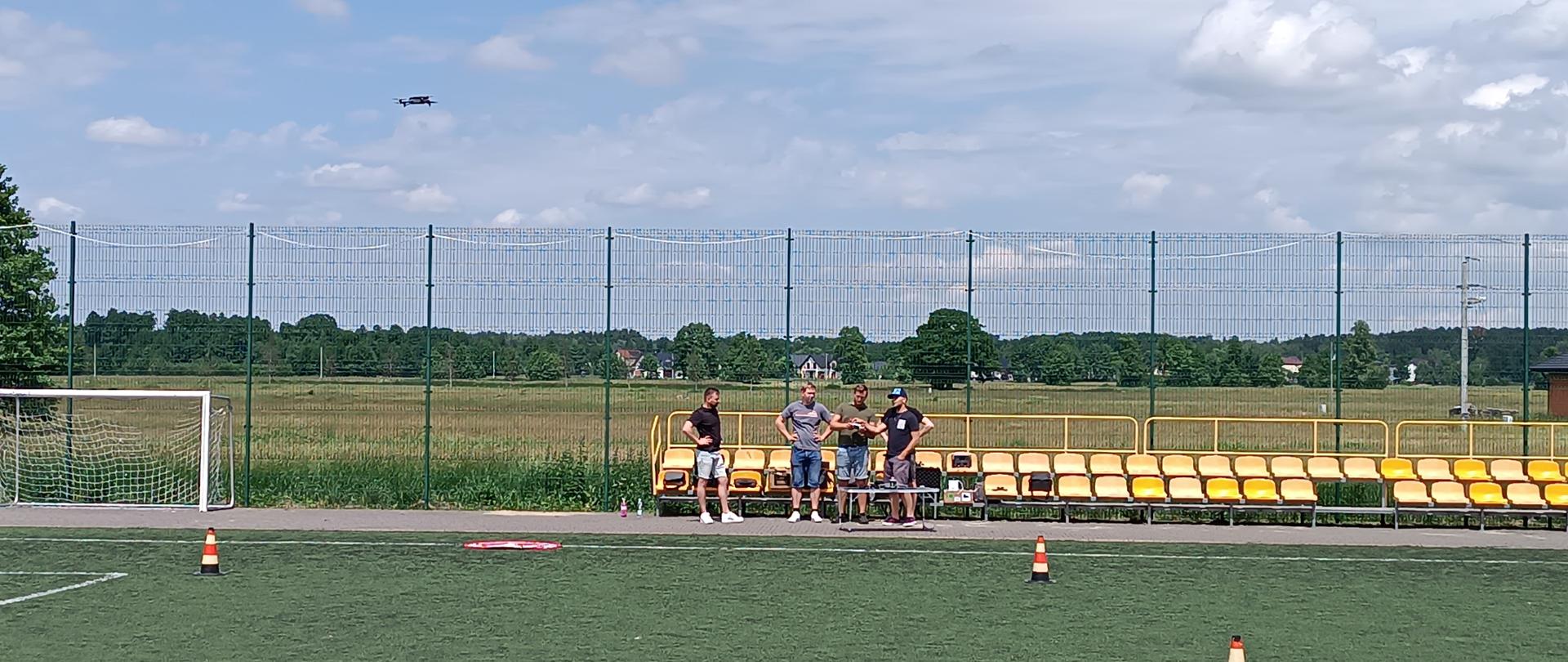 Boisko piłkarskie ze sztuczną trawą. Przy żółtych trybunach stoją trzej mężczyźni — jeden trzyma kontroler drona. W lewym górnym rogu nieba widoczny lecący dron. Na murawie rozstawione pachołki, po lewej stronie widoczna bramka