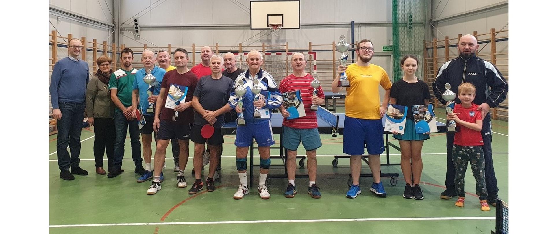Uczestnicy turnieju z przedstawicielami władz Gminy Hażlach. Na wspólnej fotografii zawodnicy w różnym wieku, niektórzy prezentują puchary i dyplomy. Z tyłu za nimi stół do tenisa stołowego.