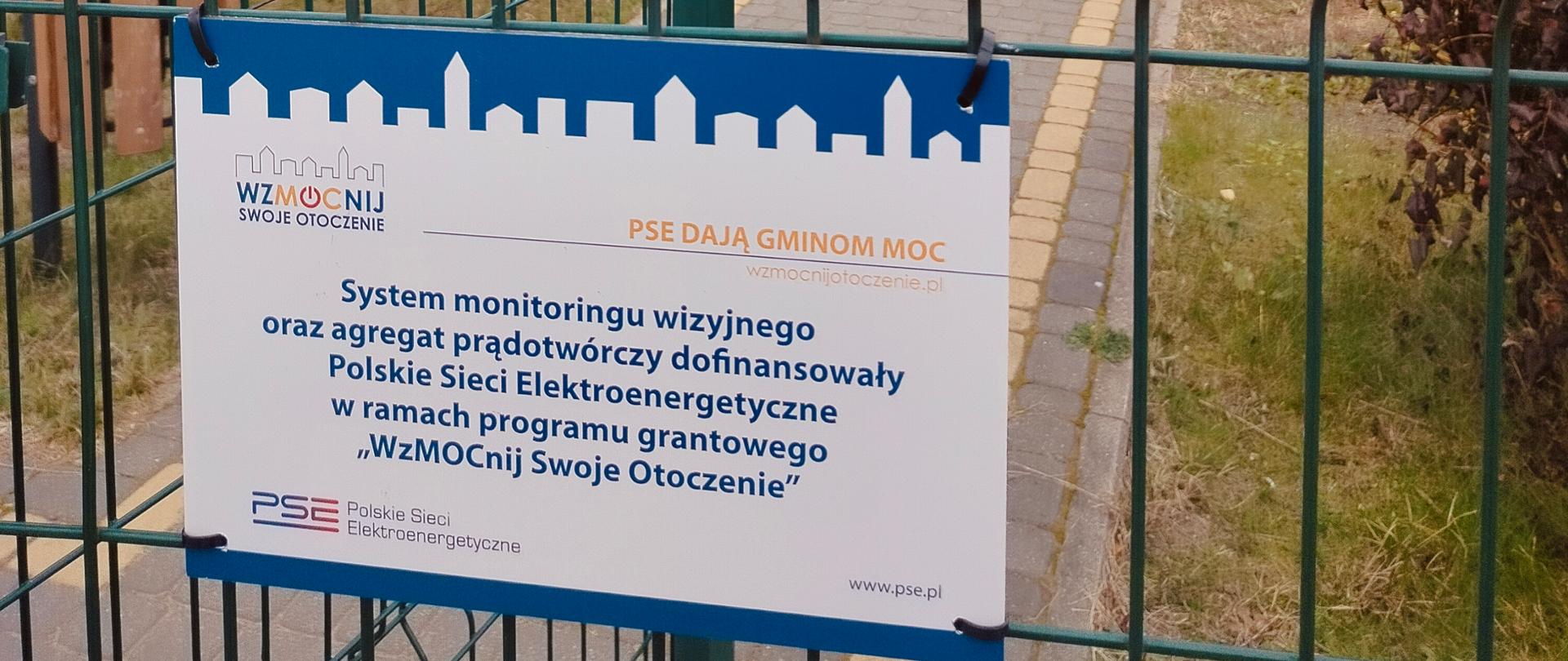 Tablica informacyjna zawieszona na zielonym ogrodzeniu panelowym. Za tablicą chodnik z kostki i parterowy budynek z lewej strony.