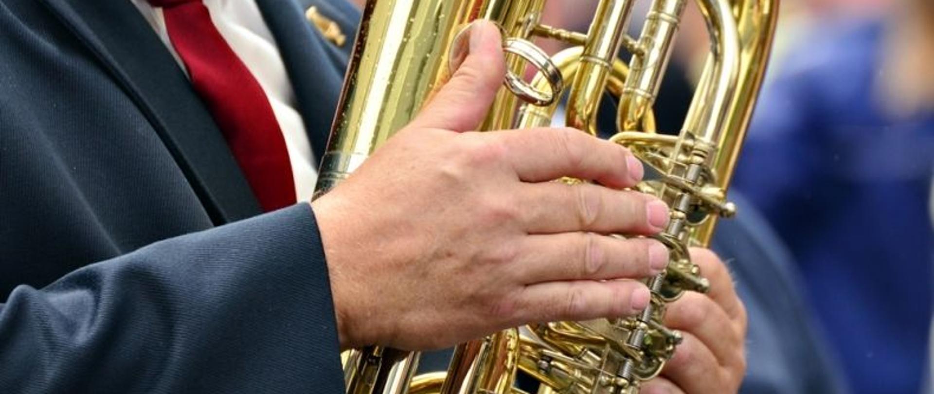 Korpus oraz dłonie muzyka który gra na trąbie.