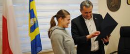 Uśmiechnięta Martyna Gacek i Wójt Gminy Raba Wyżna w chwili odbioru wyróżnienia, Wójt trzyma w ręce nagrodę. Obok postaci znajdują się dwie flagi oraz herb Gminy Raba Wyżna