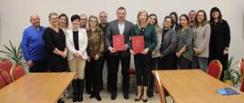 Grupa kobiet i mężczyzn ustawiona obok siebie w 2 rzędach. Na środku stoją mężczyzna i kobieta którzy trzymają oburącz przed sobą czerwone okładki na dokumenty