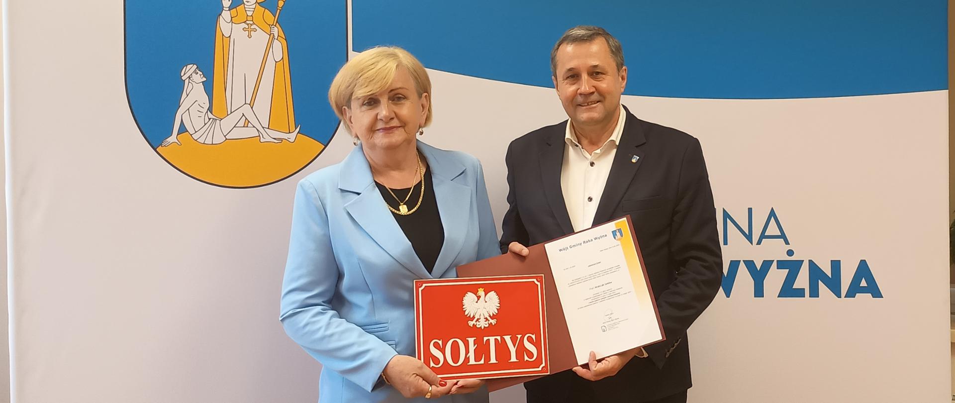 Mężczyzna w czarnym garniturze trzymający zaświadczenie o wyborze sołtysa oraz kobieta w niebieskim garniturze trzymająca tabliczkę Sołtys na tle ścianki promocyjnej z logiem Gminy Raba Wyżna.