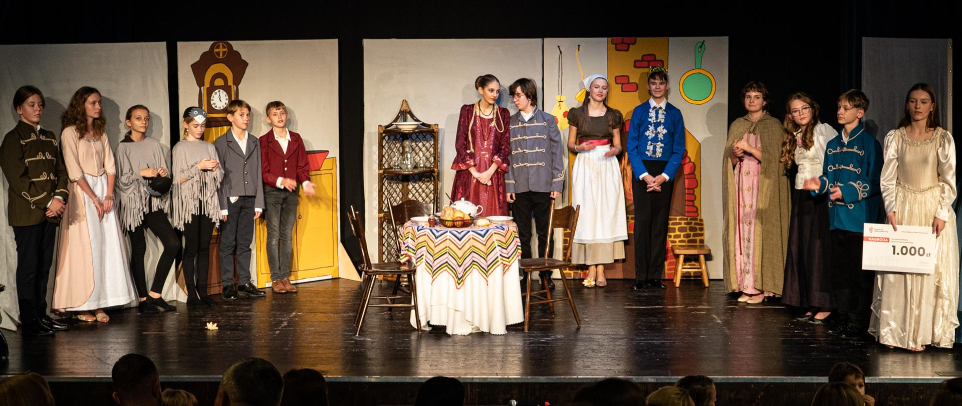 Grupa osób w kolorowych strojach, którzy przedstawiają spektakl kopciuszek, wśród nich dziewczynki-ptaki, macocha, książę, oraz główna bohaterka - Kopciuszek, która w ręku trzyma plansz- czek z kwotą 1.000 zł.