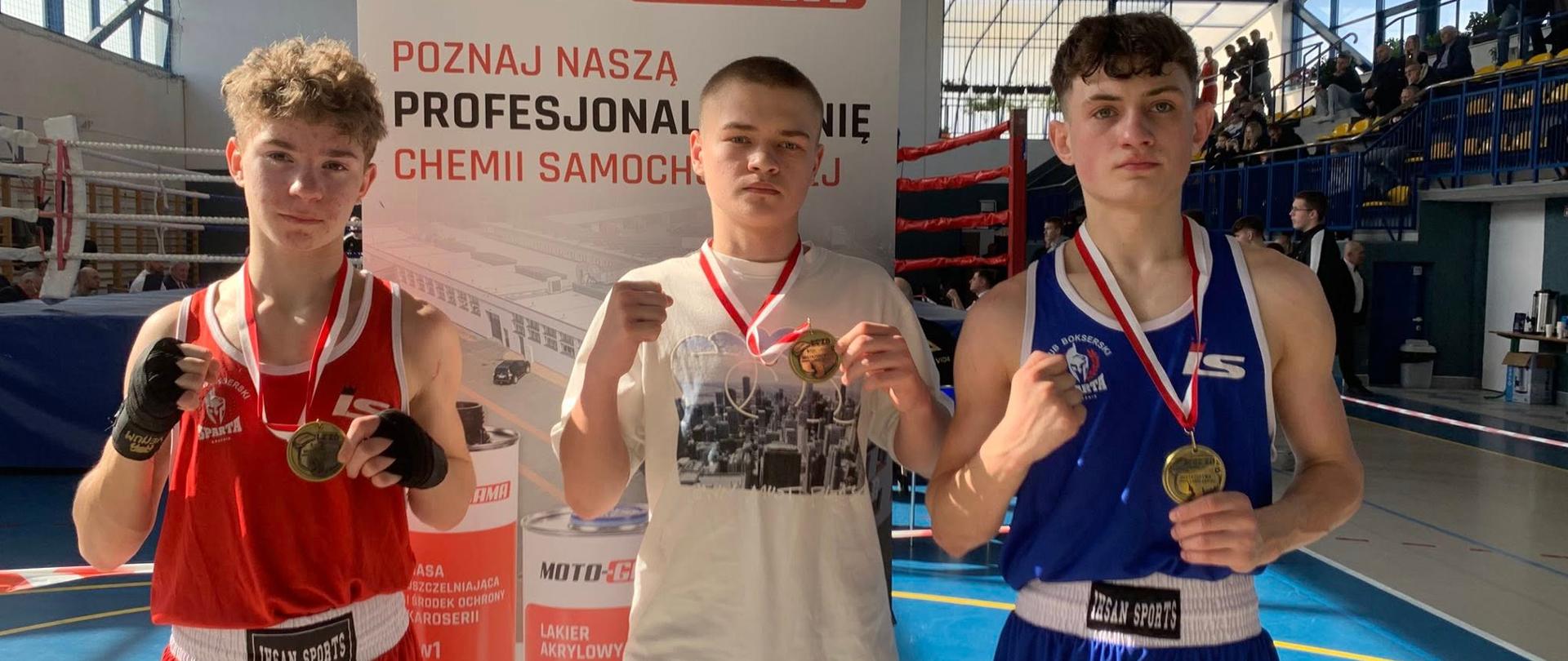 Trzech zawodników z Klubu Bokserskiego "Sparta" Kraśnik stojących obok siebie na sali sportowej prezentuje zawieszone na szyjach medale