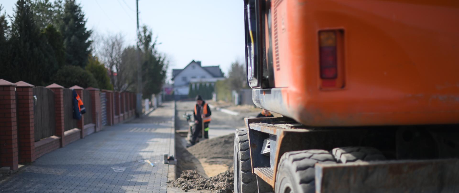 Trwają prace drogowe przy budowie chodnika na osiedlowej ulicy. W kadrze widać koparkę stojącą przy krawędzi drogi oraz pracowników w odblaskowych kamizelkach wykonujących roboty ziemne. Po lewej stronie znajduje się ułożony fragment nowego chodnika z kostki brukowej, a w tle widoczne są ogrodzenia i domy jednorodzinne.