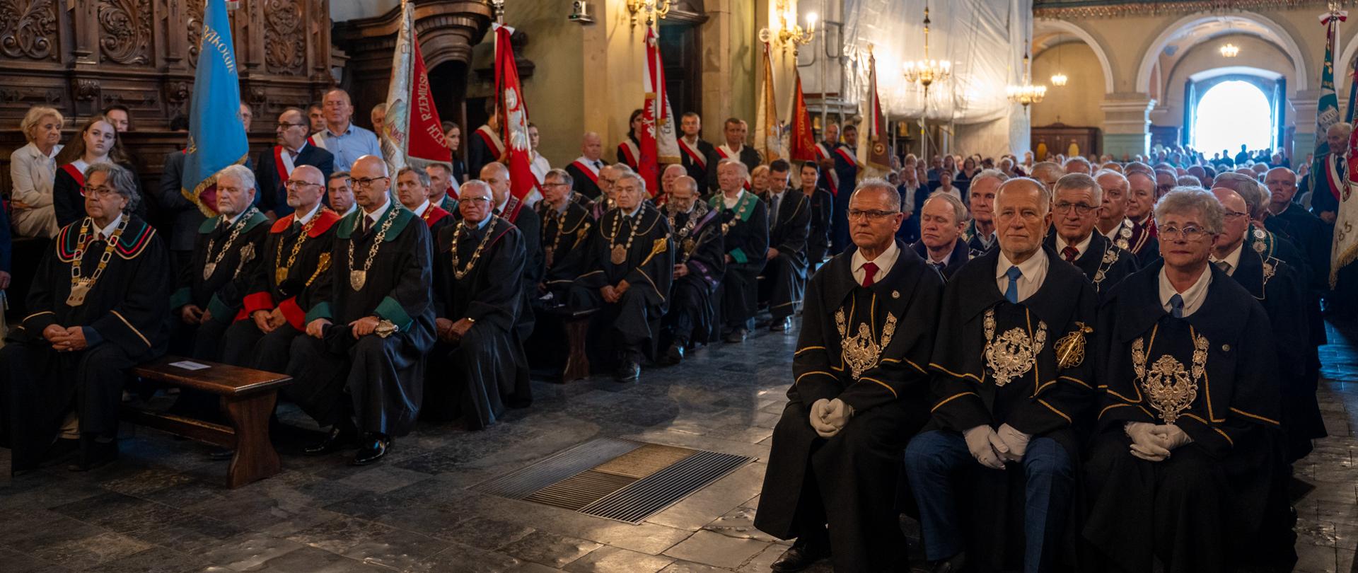 Uczestnicy Małopolskiej Pielgrzymki Rzemieślników i Kupców zgromadzeni podczas nabożeństwa w bazylice w Kalwarii Zebrzydowskiej. W pierwszych rzędach siedzą przedstawiciele cechów w strojach organizacyjnych z insygniami, a w tle widoczne są poczty sztandarowe oraz licznie zgromadzeni wierni.
