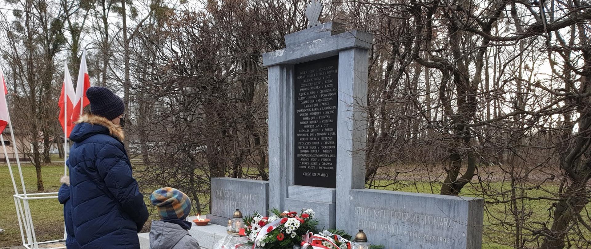 Dorosły z dwójką chłopców stoi przed pomnikiem. Leżą przed nim wiązanki kwiatów, płoną znicze.