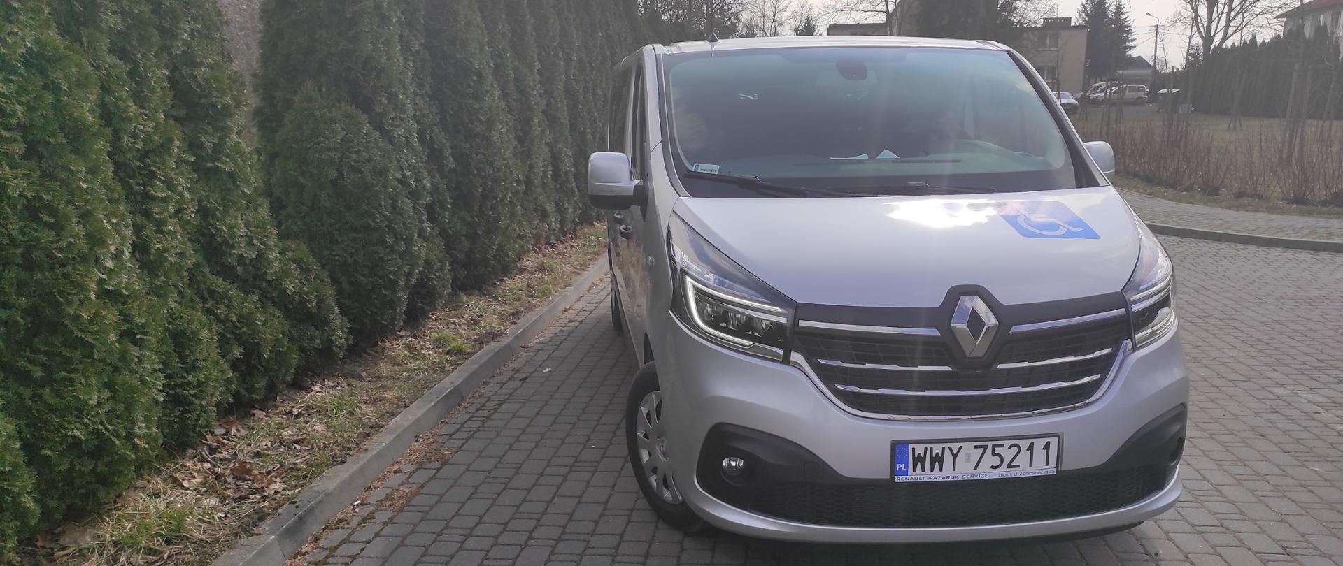 Srebrny samochód Renault Trafic ustawiony przodem na szarej kostce. Po lewej stronie zdjęcia wysokie zielone thuje