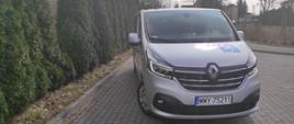Srebrny samochód Renault Trafic ustawiony przodem na szarej kostce. Po lewej stronie zdjęcia wysokie zielone thuje