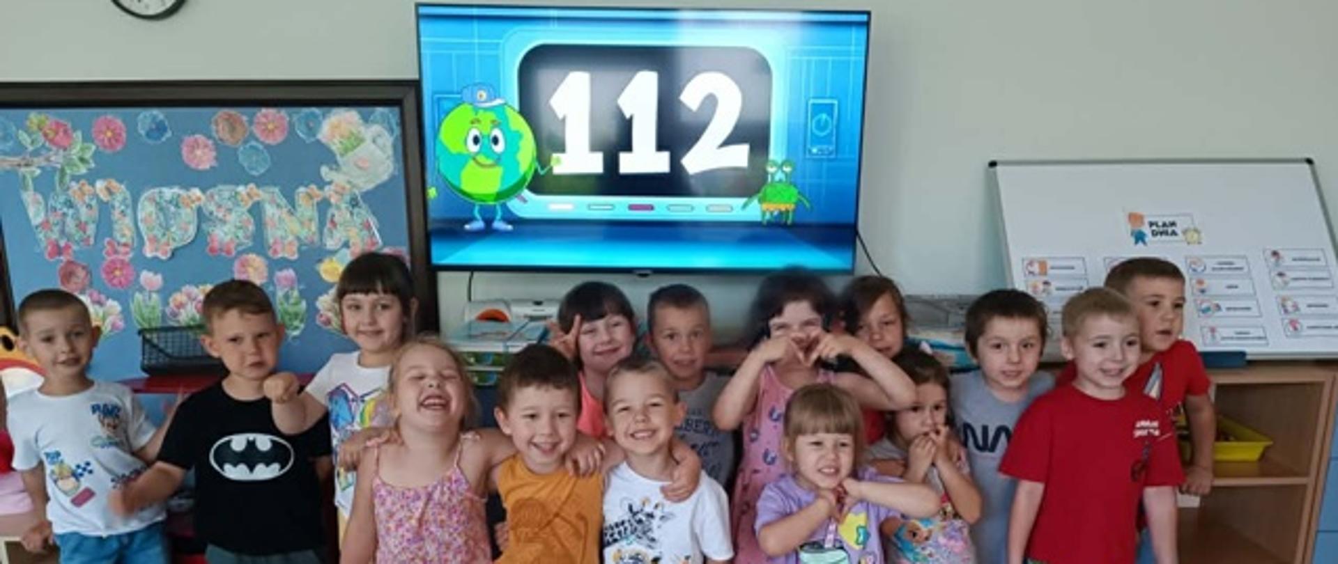 Dzieci pozują do zdjęcia, za nimi monitor z numerem alarmowym 112