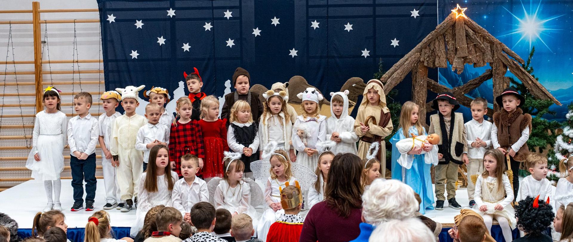 Na scenie ustawionej w sali gimnastycznej występuje grupa najmłodszych dzieci w świątecznych strojach. Mali artyści przebrani są m.in. za aniołki, pasterzy, owieczki oraz postacie z bożonarodzeniowej szopki. W tle widoczna jest dekoracja przedstawiająca nocne niebo pełne gwiazd oraz stajenkę z betlejemską gwiazdą. Przed sceną zgromadzona jest publiczność, w tym rodzice i bliscy dzieci, uważnie obserwujący występ.