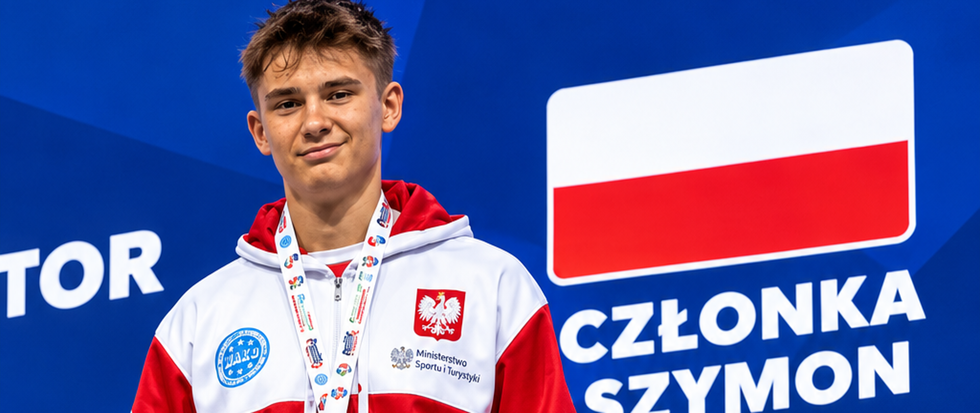 Szymon Członka stojący na niebieskim tle z flagą Polski, pod którą jest napis: Szymon Członka Poland, a poniżej grafika brązowego medalu. Szymon ubrany jest w biało - czerwony dres, na szyi ma zawieszony brązowy medal, a w rękach trzyma dyplom.