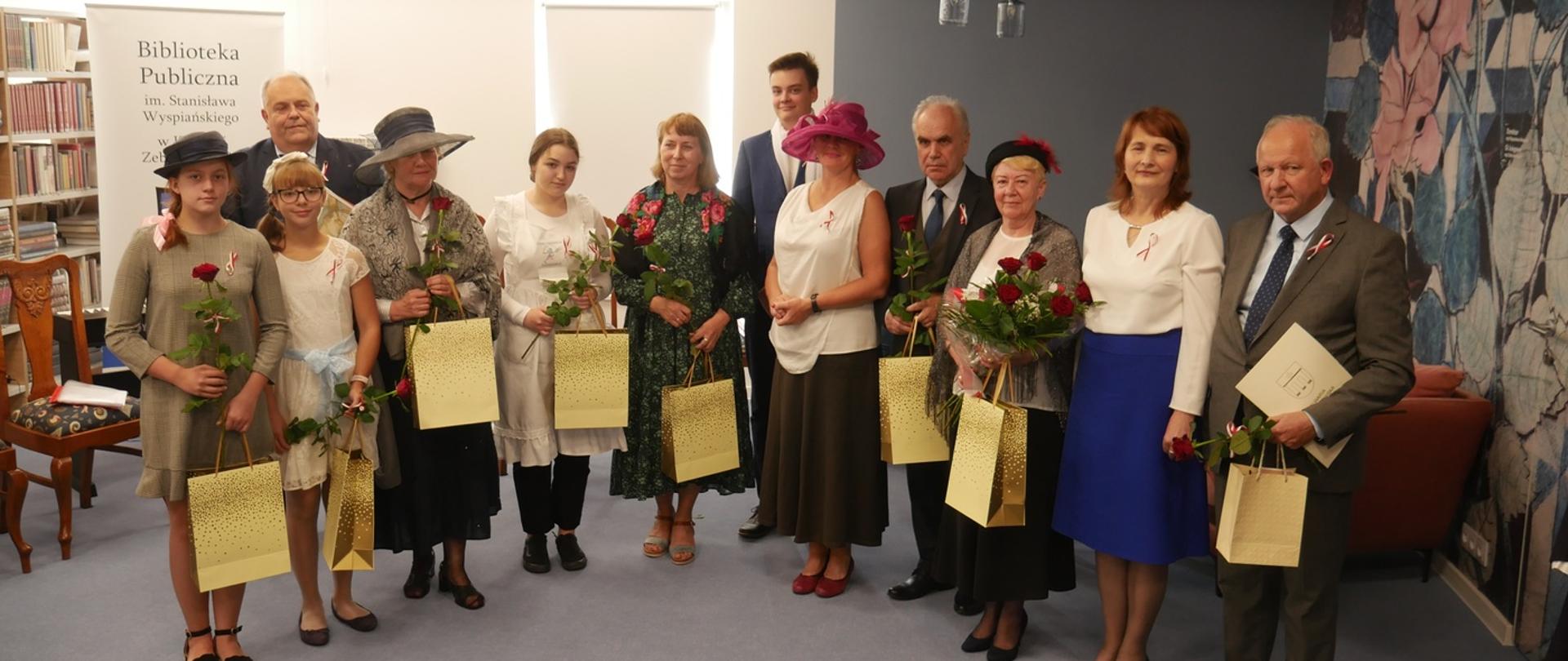 Uczestnicy Narodowego Czytania pozujący do grupowego zdjęcia w bibliotecznej czytelni. Lektorzy mają na sobie stylizowane stroje, w rękach trzymają kwiaty i torebki upominkowe.