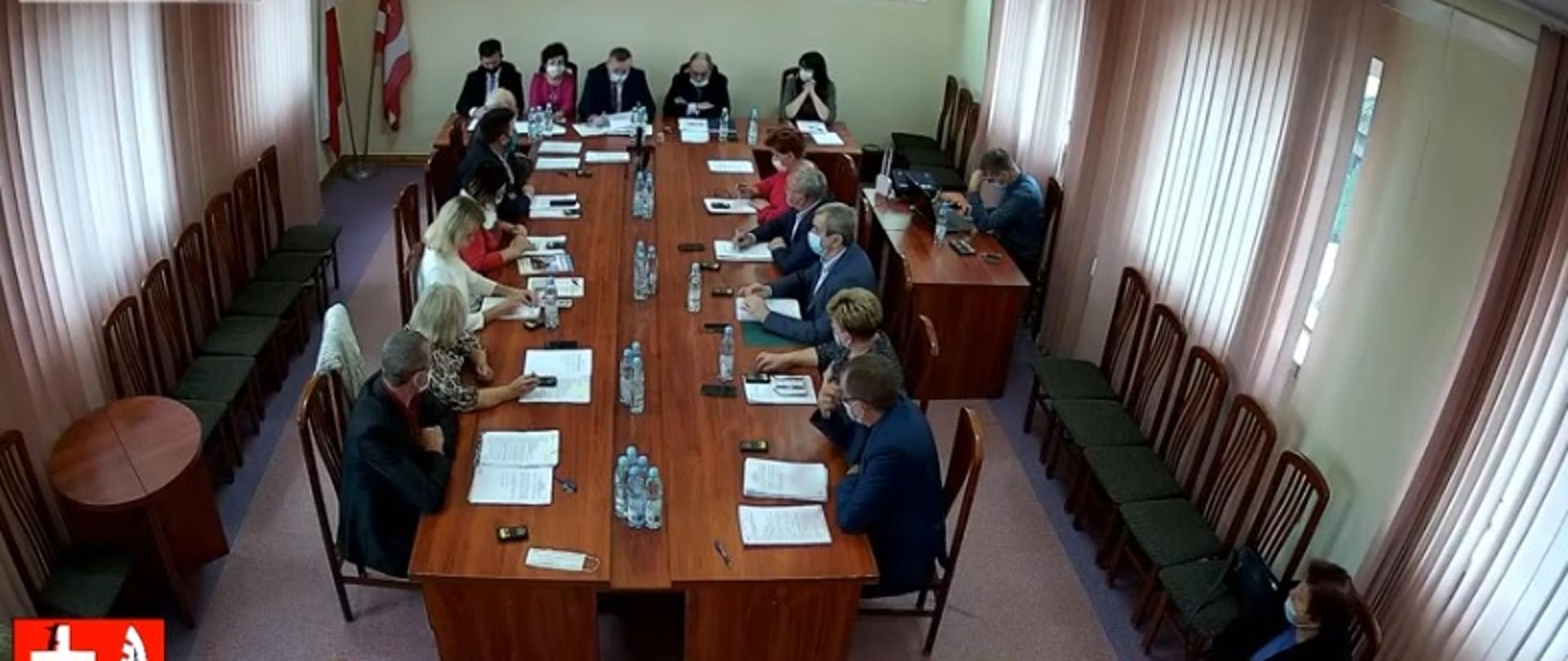 Przy stole Radni Gminy Zabrodzie, Wójt, Skarbnik, Sekretarz, na stole kartki, woda, na ścianie godło Polski, herb, ekran projektora, na dole ekranu herb Gminy Zabrodzie