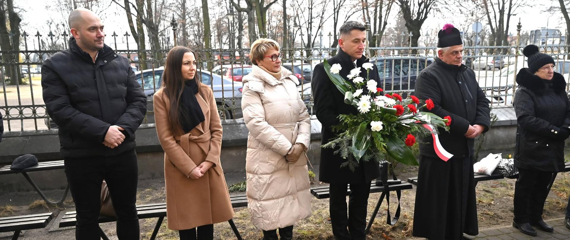 Władze miasta z wiązankami kwiatów pod tablicą.