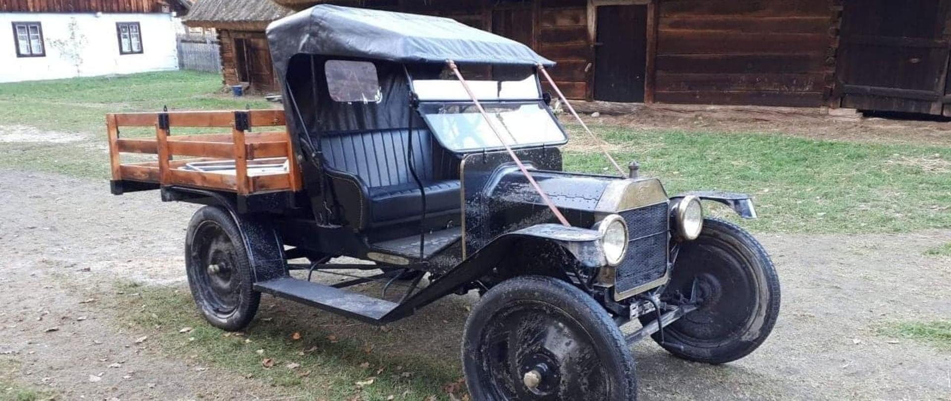 Na zdjęciu widać epokowy samochód marki Ford z 1920 roku w kolorze czarnym. W tle po lewej stronie widać starszy wiejski dom, a obok stoi wielka drewniana szopa.