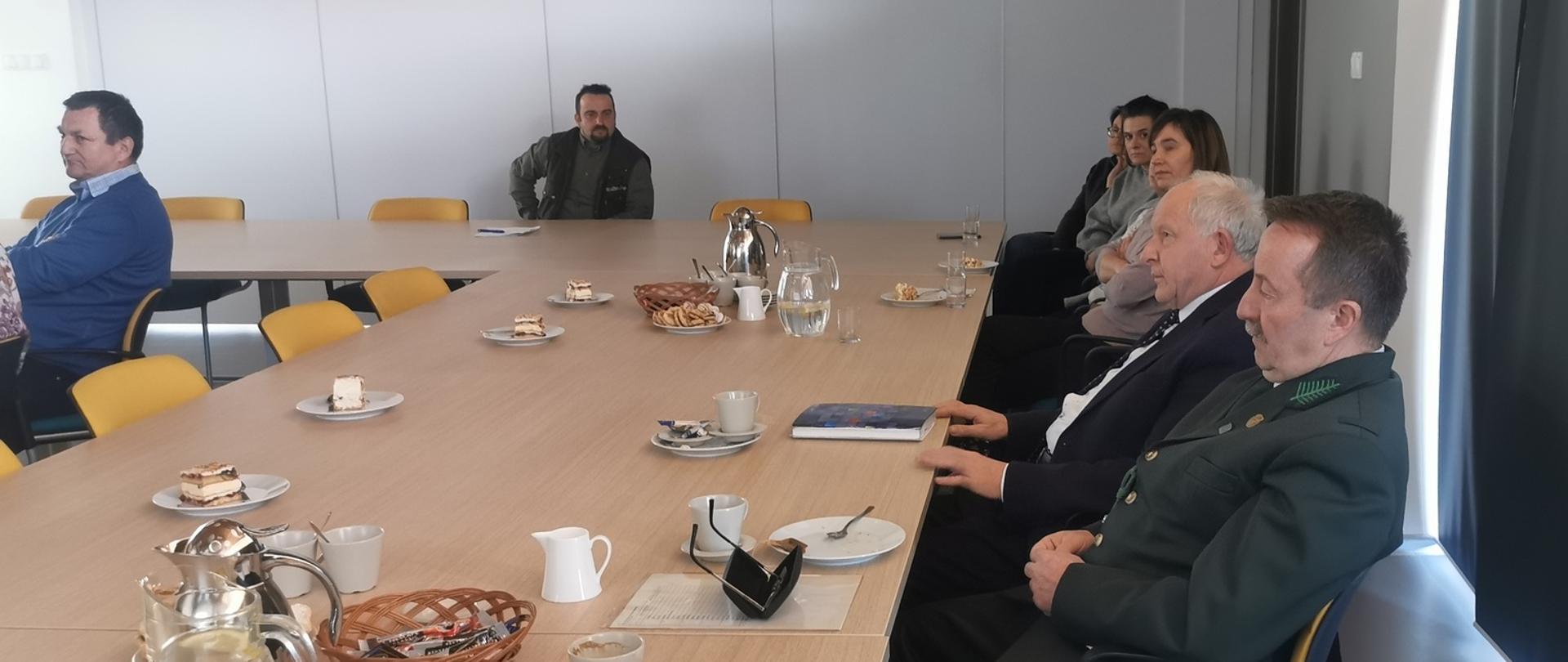 Uczestnicy spotkania siedzą przy dużym stole, na stole filiżanki, dzbanki, talerzyki, koszyki z kawą, notatniki.