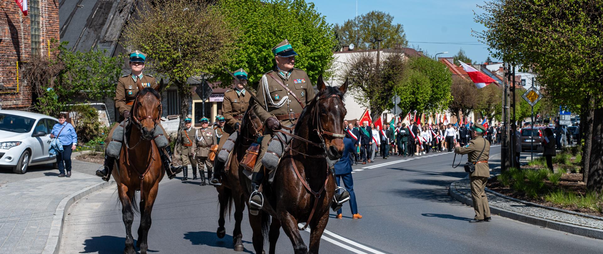 Uroczysty przemarsz ulicą miasta podczas obchodów Święta Konstytucji 3 Maja – na pierwszym planie trzej umundurowani jeźdźcy na koniach, za nimi długa kolumna uczestników niosących biało-czerwone flagi i sztandary; słoneczny dzień, zabudowa miejska i drzewa w tle.