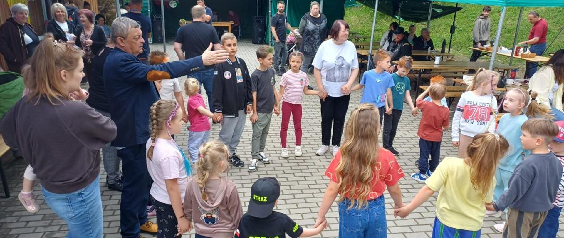 Grupa dzieci i dorosłych uczestniczy w pikniku rodzinnym na placu przed Domem Ludowym. Uczestnicy stoją w dużym kręgu, trzymając się za ręce podczas wspólnej zabawy integracyjnej. W tle widoczne są namioty, stoły biesiadne oraz mieszkańcy spędzający czas w rodzinnej i radosnej atmosferze.