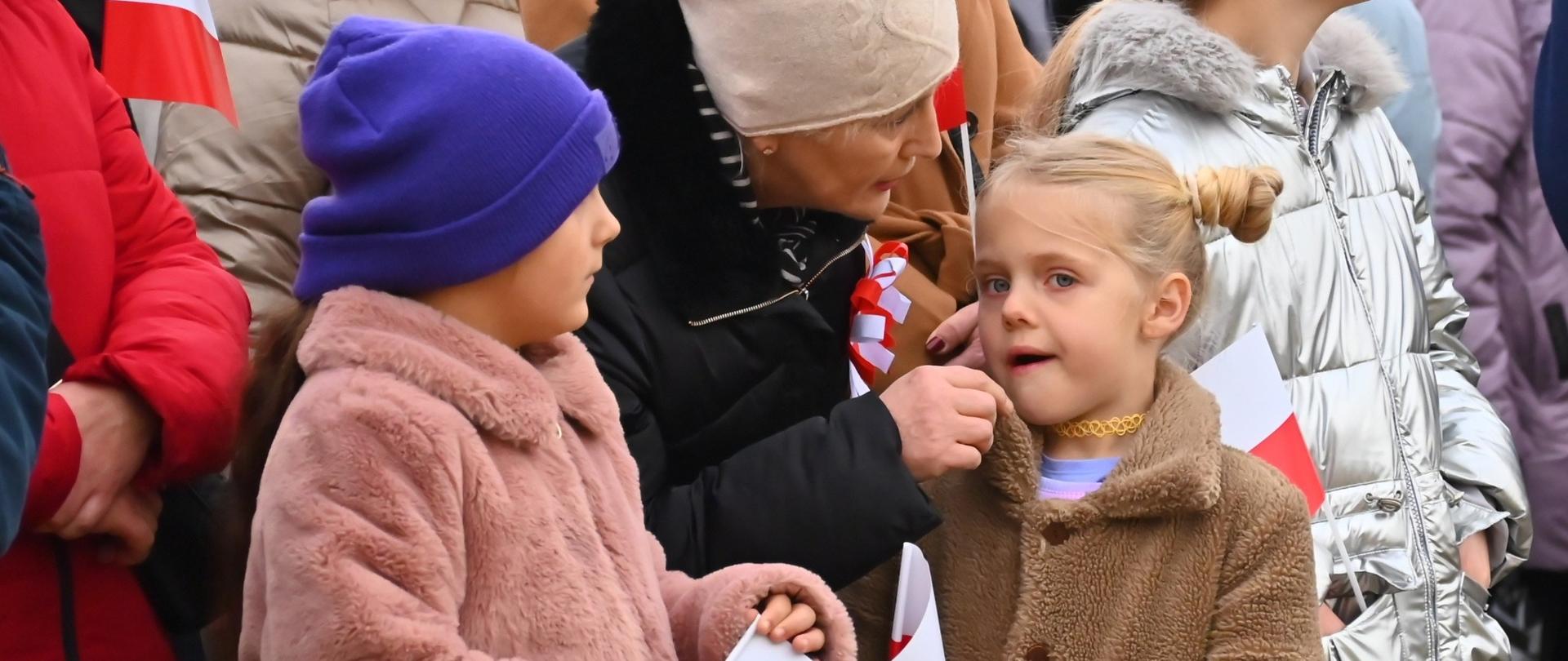 Dzieci w kolorowych czapkach i ciepłych kurtkach trzymają biało-czerwone flagi. 