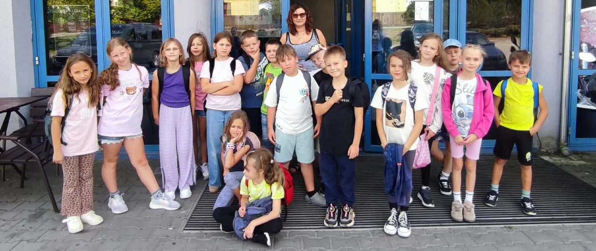 Grupa dzieci wraz z opiekunką pozuje do wspólnego zdjęcia przed wejściem do budynku z dużymi przeszkleniami i niebieskimi drzwiami. Dzieci ubrane są w letnie stroje, a część z nich ma ze sobą plecaki.