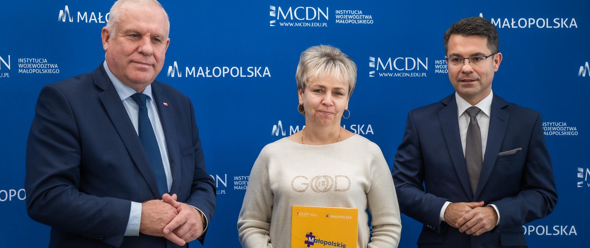 Na fotografii widnieją trzy osoby stojące pośrodku kobieta blondynka trzymająca przed sobą kolorową teczkę po jej prawej i lewej stronie mężczyźni w garniturcach