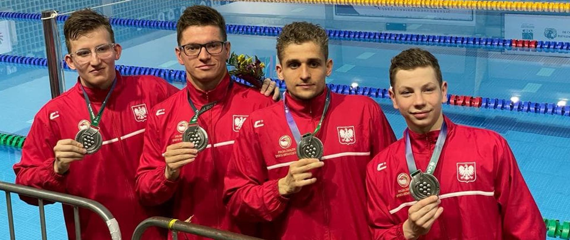 Zdjęcie przedstawia 4-osobową sztafetę 4 x 200 m stylem dowolnym, stojącą na tle brazylijskiego basenu. Zawodnicy ubrani są w stroje reprezentacji Polski i prezentują w dłoniach zawieszone na szyjach srebrne medale. Pierwszy od prawej strony stoi Rafał Wójcik.