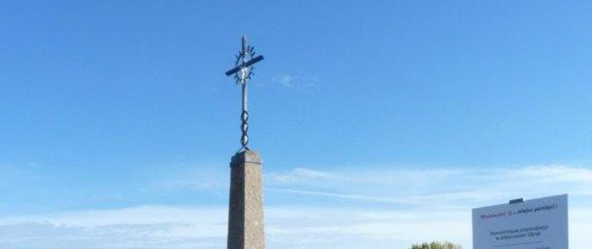 Krzyż stalowy na betonowej wąskiej wysokiej podstawie. Wokół niskie metalowe ogrodzenie