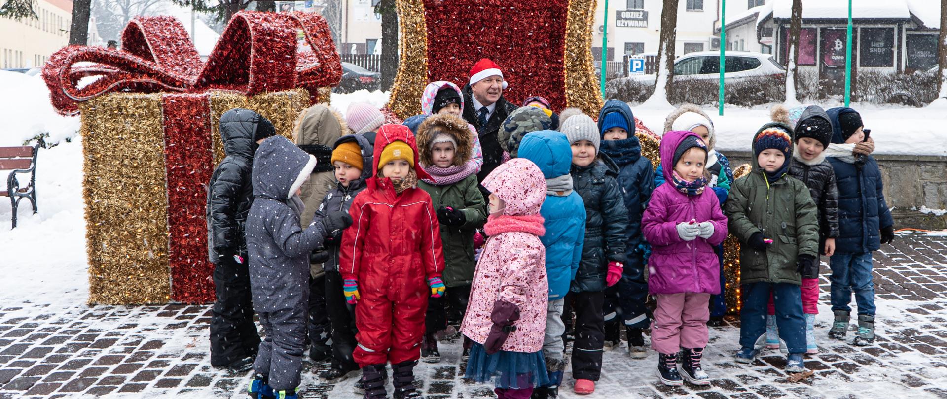 W tle fotel św. Mikołaja oraz prezent zrobiony ze światełek w kolorach czerwonych i żółtych. Na fotelu siedzi mężczyzna w czapce mikołaja, wokół niego jest zebrana gromadka dzieci.