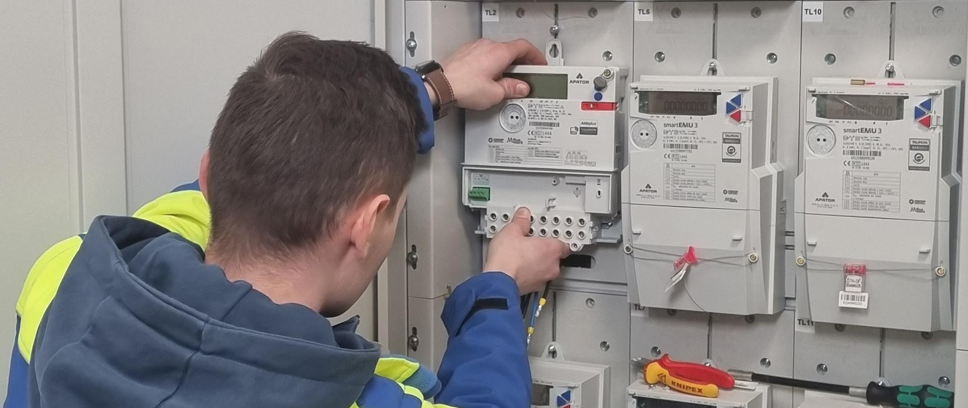 Młody elektryk w niebiesko-żółtej bluzie montuje licznik energii w szafie montażowej