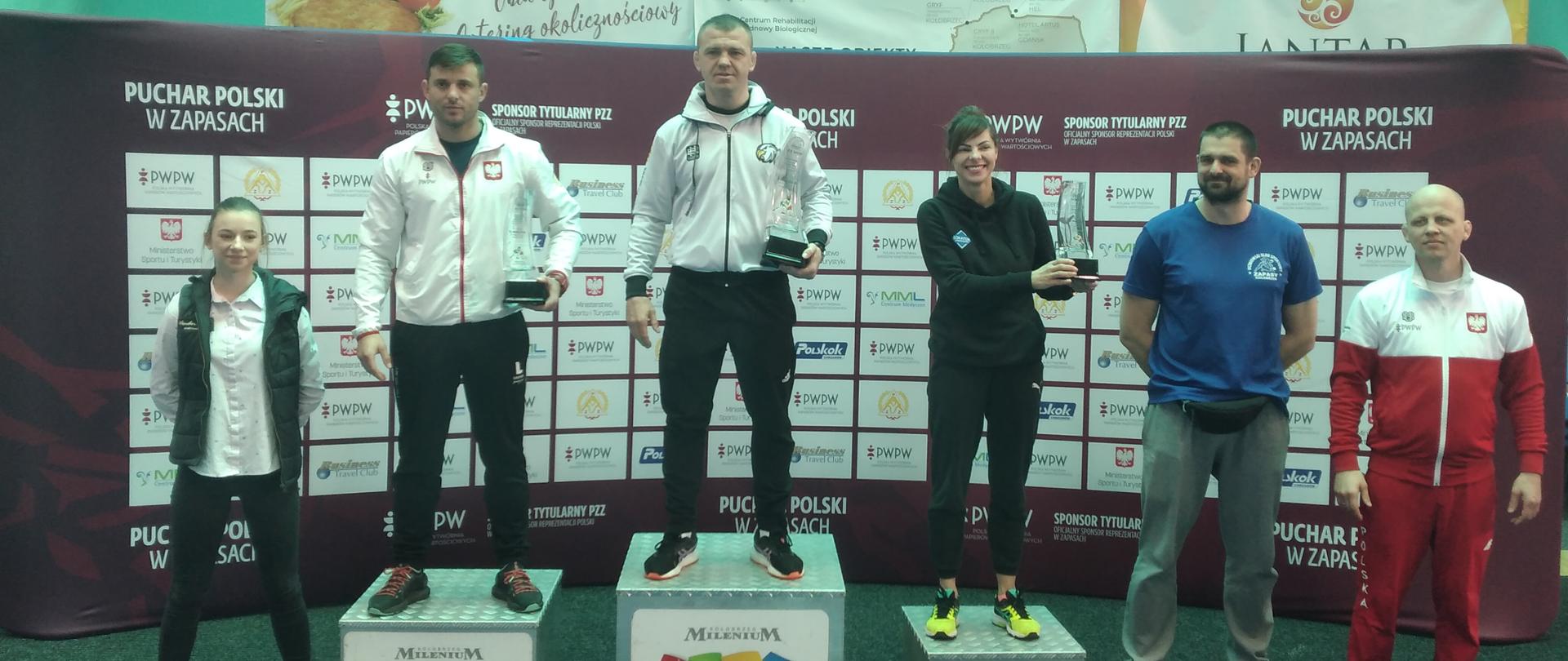 Zdjęcie przedstawia trzech trenerów drużyn zapaśniczych na podium rywalizacji drużynowej II Pucharu Polski Juniorów i Juniorek młodszych w zapasach styl wolny i zapasach kobiet. Na drugim stopniu stoi trener Ludowego Uczniowskiego Klubu Sportowego "Suples" w Kraśniku Damian Jakóbczyk. Trenerzy trzymają w rękach puchary