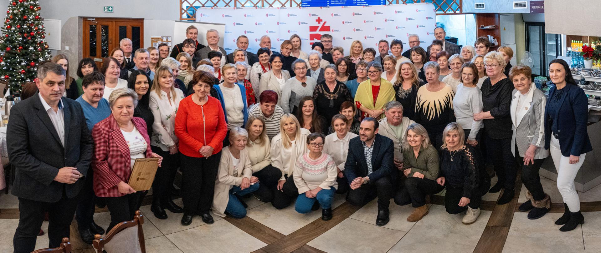 Grupowe zdjęcie uczestników spotkania integracyjnego w klasztorze w Kalwarii Zebrzydowskiej. Kilkadziesiąt osób – głównie dorosłych i seniorów – pozuje we wnętrzu sali, ustawionych w kilku rzędach. W tle widoczna jest choinka bożonarodzeniowa oraz ścianka z logo wydarzenia. Uczestnicy ubrani są w eleganckie i półformalne stroje, część osób siedzi w pierwszym rzędzie, pozostali stoją. Zdjęcie dokumentuje wspólne, uroczyste spotkanie środowisk lokalnych.