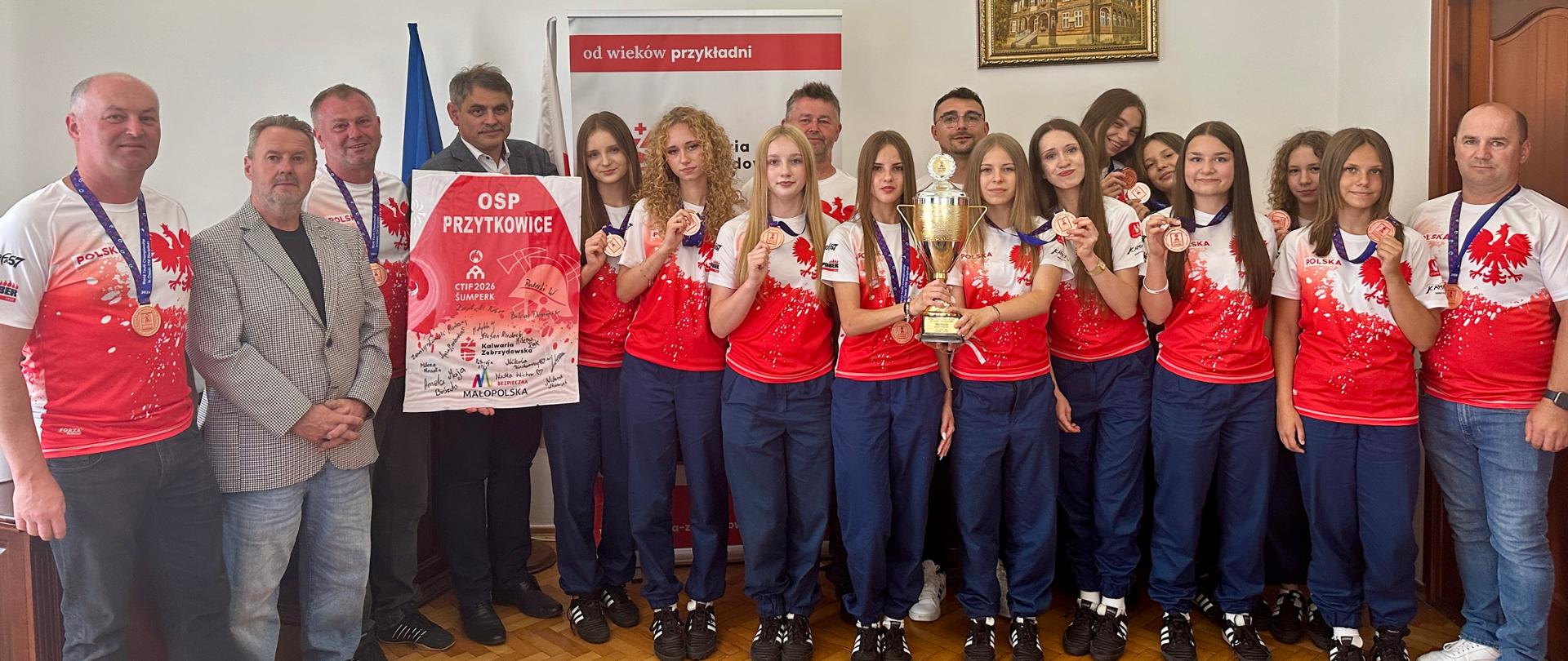Wspólne zdjęcie zawodniczek Młodzieżowej Drużyny Pożarniczej Dziewcząt OSP Przytkowice, trenerów oraz przedstawicieli samorządu podczas spotkania w Urzędzie Miasta Kalwarii Zebrzydowskiej. Drużyna prezentuje brązowe medale oraz puchar zdobyty podczas Międzynarodowej Olimpiady Młodzieżowych Drużyn Pożarniczych CTIF w czeskim Šumperku.