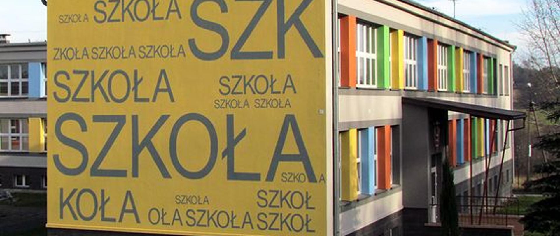 Budynek szkoły z kolorową elewacją pomiędzy oknami, bok budynku ozdobiony różnej wielkości napisami "szkoła".
