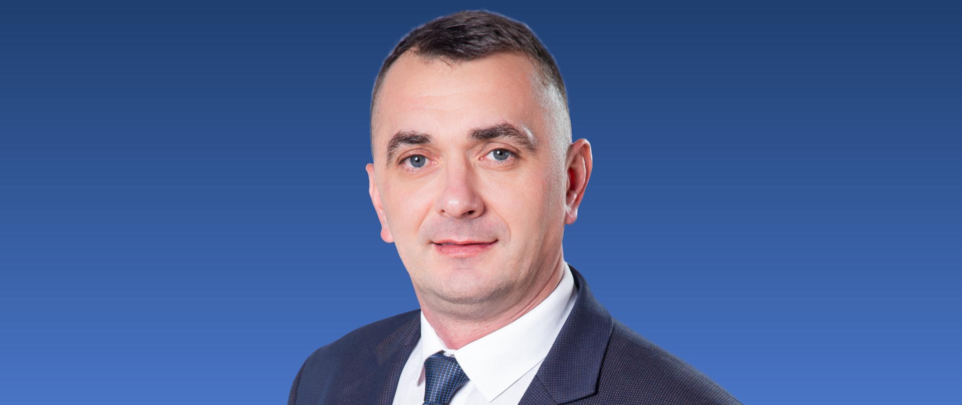 Zdjęcie przedstawia mężczyznę w krawacie na niebieskim tle.