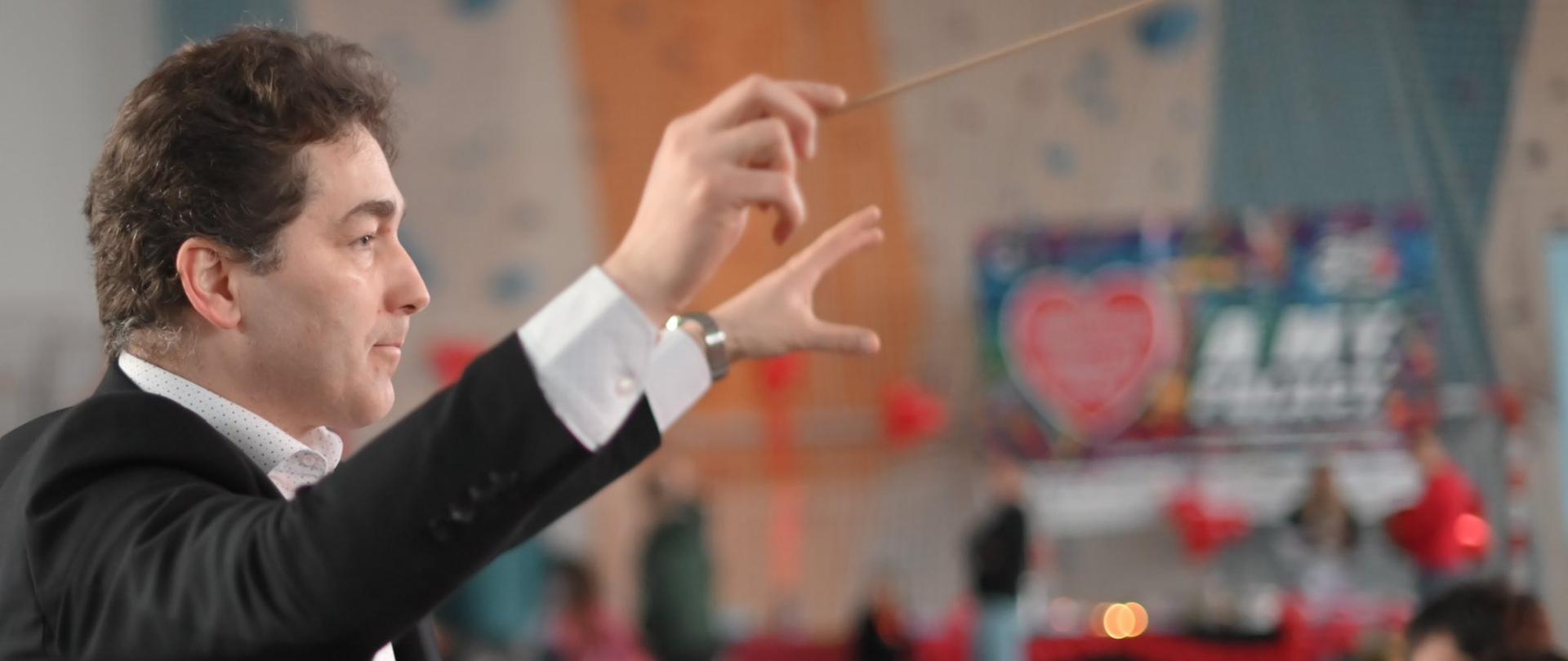 Dyrygent prowadzący orkiestrę podczas koncertu w hali – mężczyzna w garniturze, z uniesionymi rękami i batutą, w tle muzycy oraz dekoracje wydarzenia.